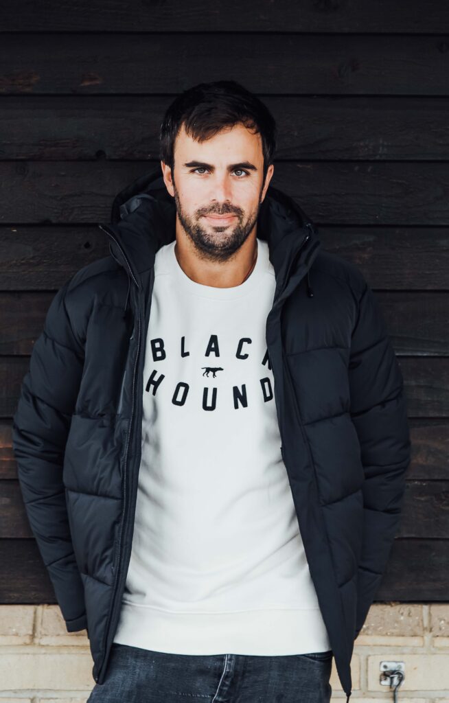 Branding image of a man leaning against a wooden stable wearing top and coat for a sports brand.