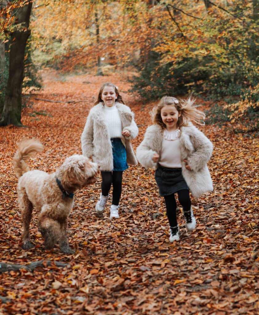2 girls in fur coats running with their dog through a woodland in an Autumnal season.