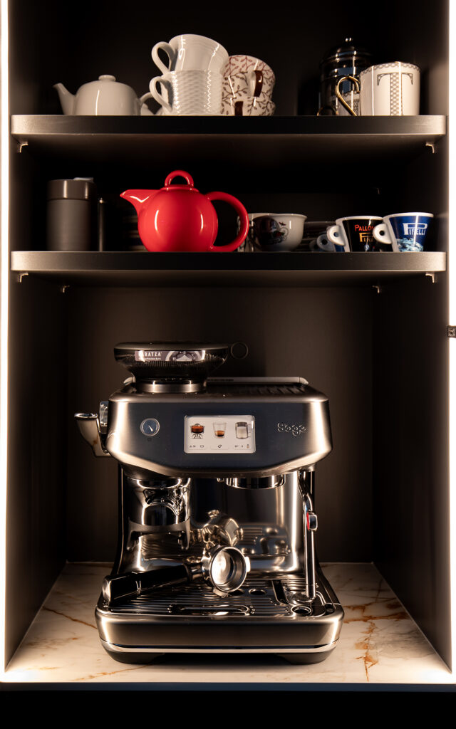 Styled photo of a coffee machine inside a cupboard with a red teapot and white china