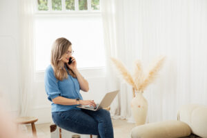 Lady sitting in a blue shirt and jeans on her phone looking at her laptop