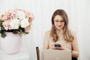 Light and airy studio shot of a lady looking down at her phone with laptop in front of here.